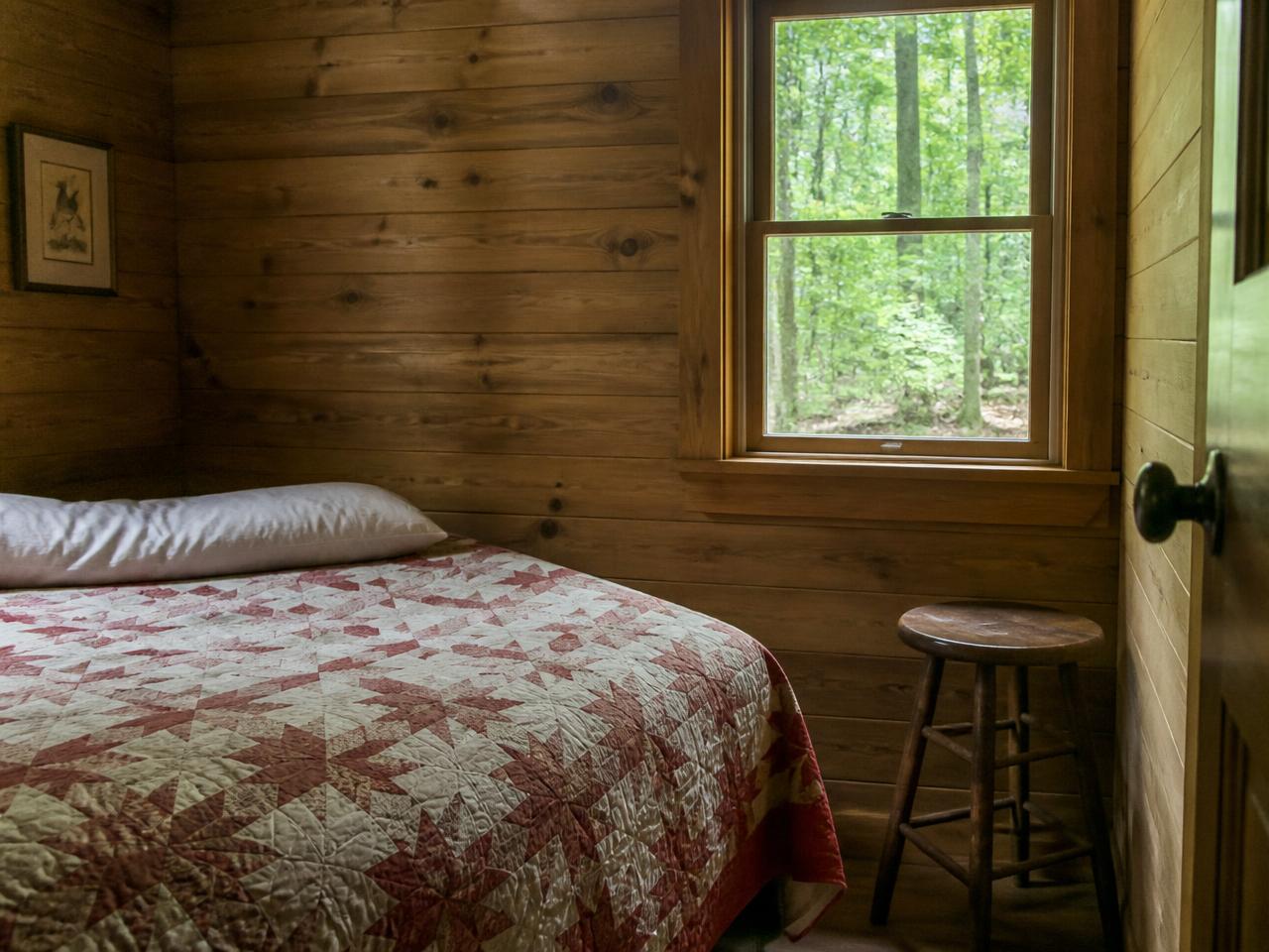 Inside a simple cabin: wood walls, a quilt-covered bed, a stool, and a window to the woods